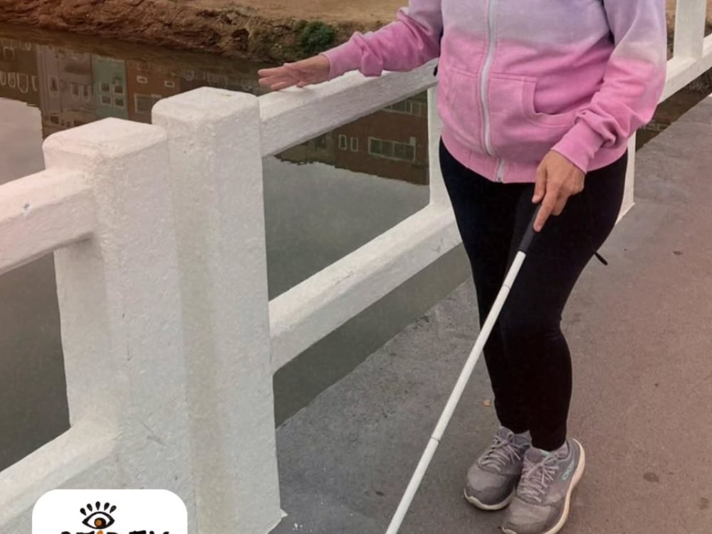 Aluna caminhando com a bengala branca ao longo de uma ponte na cidade, em aula de orientação e mobilidade