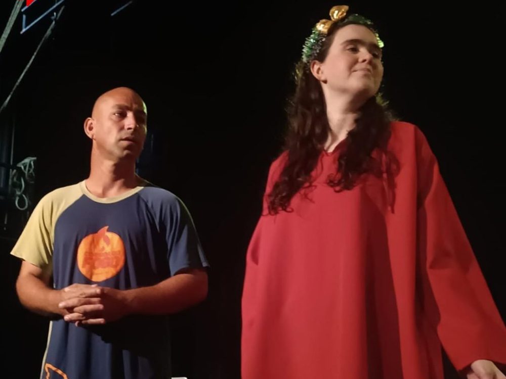 Dois atores da Cia de Teatro Novo Olhar em cena, de túnica vermelha e coroa de flores, no espetáculo A História Não Acaba Aqui