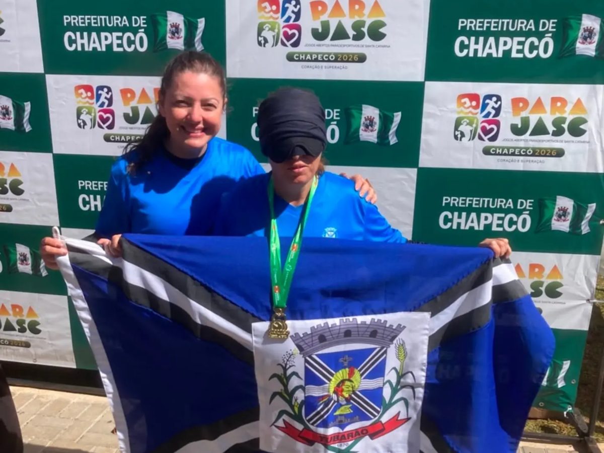 Sandra, de agasalho azul da delegação, segura a bandeira de Tubarão com a medalha no peito, no pódio do Parajasc 2026